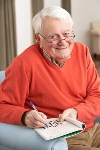 an elderly man in an orange shirt smiles as he holds a pen to a crossword puzzle book