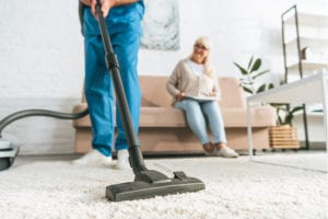 Man in scrubs vacuuming senior woman’s rug while she sits on couch and smiles.