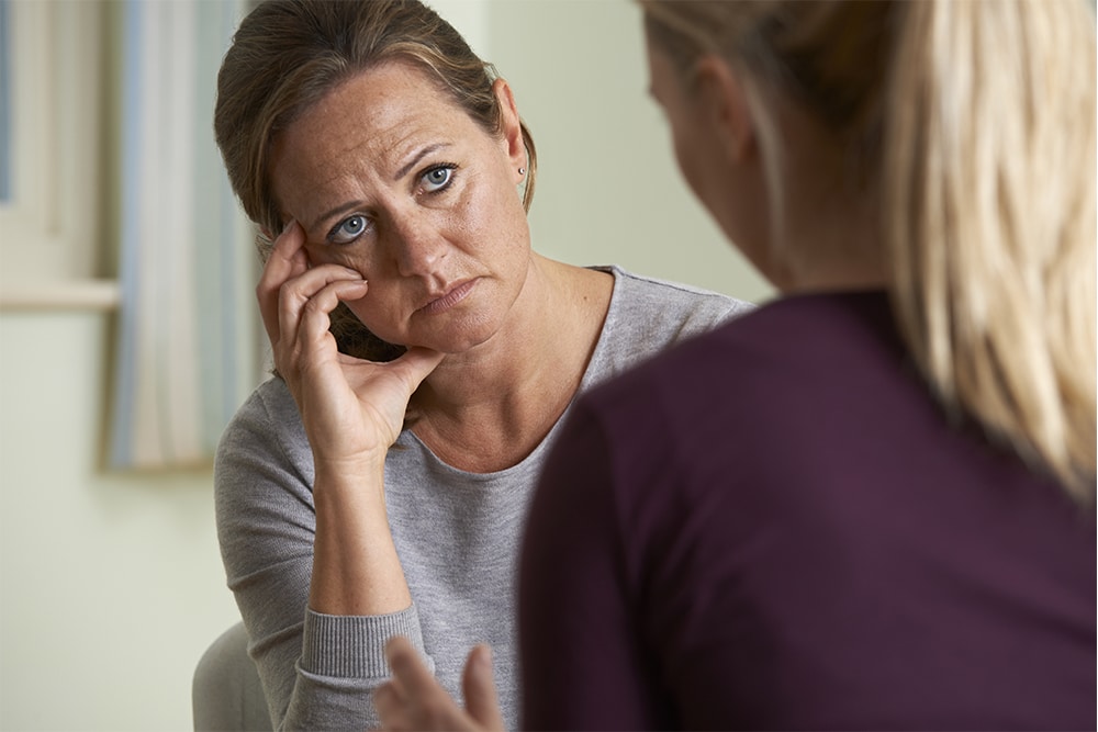 Distressed woman sits in chair looking at another woman.