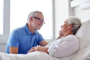 Man comforting elder woman in hospital bed.