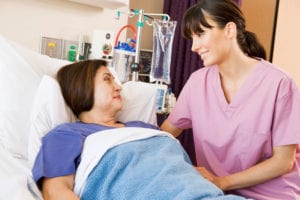 Female nurse sits to the left of female patient lying in a hospital bed