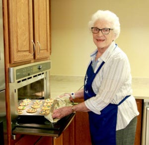A senior woman smiles at the camera as she pulls a pan of cookies out of the oven