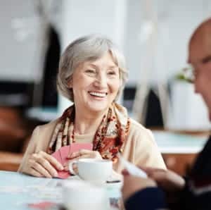 A smiling senior woman is sitting at a table playing cards and having coffee in a senior living community activity room with other senior residents.