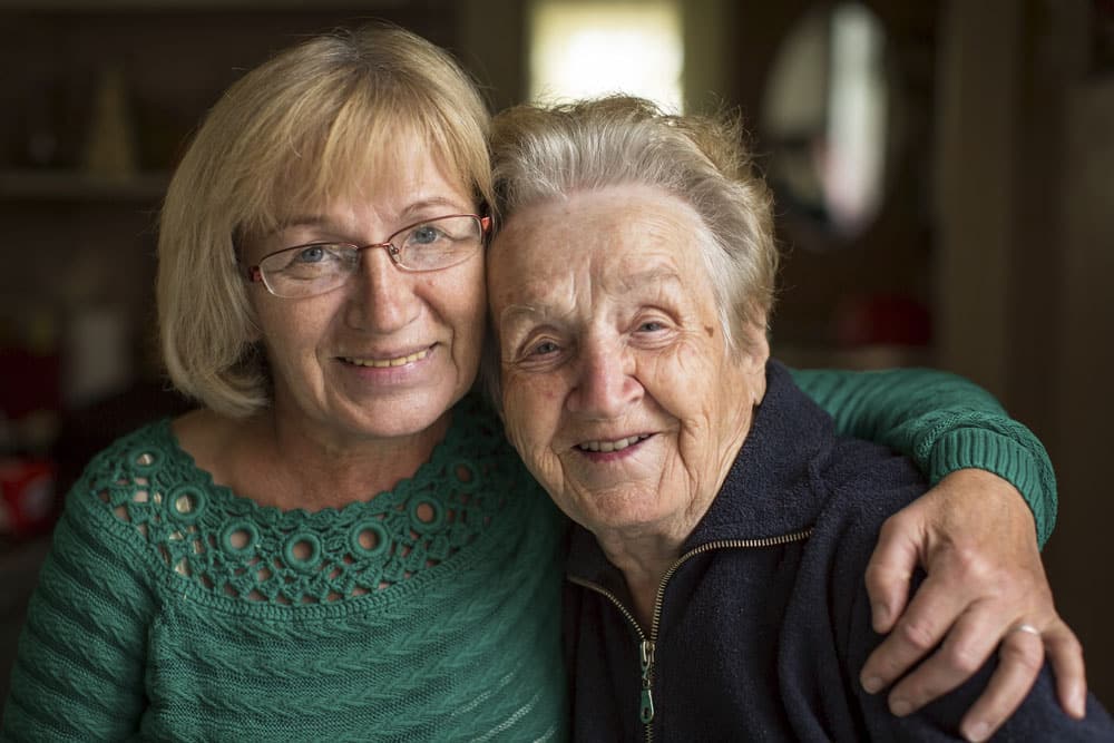 A senior woman and adult daughter are smiling and leaning in close, the daughter has her arm around her mother’s shoulder.