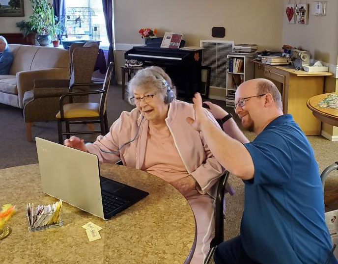 A senior woman and a male Edgewood Healthcare staff member smile and wave at a laptop during a telehealth appointment