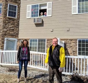 A senior couple living at Edgewood Healthcare looks out the window to pose for a photo with their son and a violinist, who are standing outside