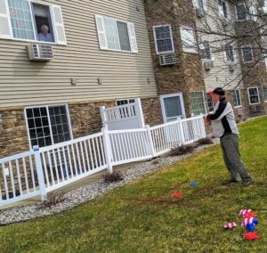 A senior female resident at Edgewood Healthcare peers out her second-story window to play tic-tac-toe with her son, who is standing near the spray-painted board on the lawn below