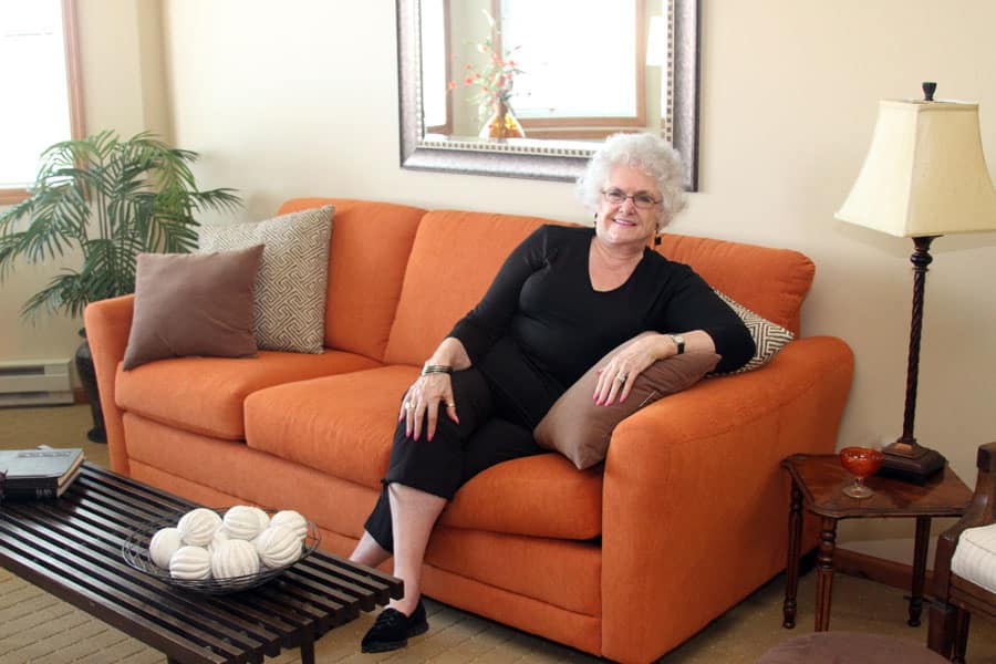 A senior woman smiles while sitting on an orange couch in a living room