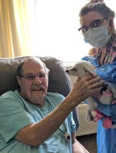 Edgewood resident Dave Bryant sits in a recliner and pets a small white dog that a female Edgewood nurse is holding in her arms on the right of the photo.