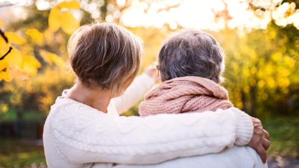 Two senior women with their backs to the camera embrace while looking at trees in autumn