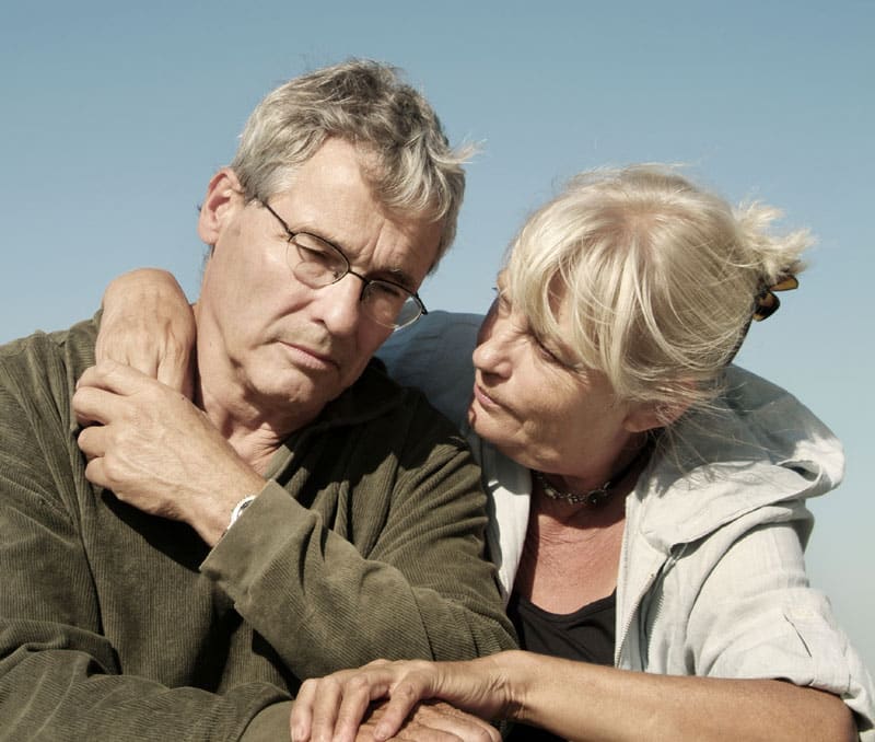 A senior woman puts her arm around a senior man who is looking down with a somber expression on his face.