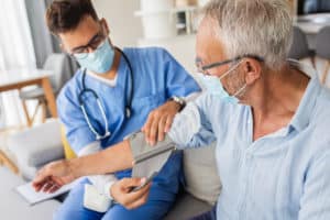 A young male doctor wearing a mask and scrubs takes a senior man’s blood pressure during an in-home health visit.