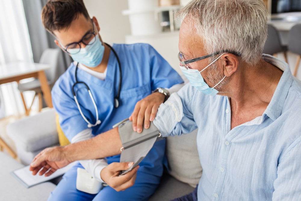 A young male doctor wearing a mask and scrubs takes a senior man’s blood pressure during an in-home health visit.