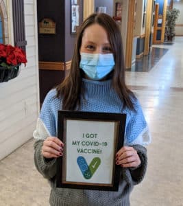 Female Edgewood Healthcare employee Kelsey Schaunaman wearing a mask and holding a framed sign that says, “I got my COVID-19 vaccine!”