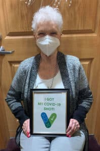 Female senior resident Hermelle Wilson sitting in a chair and holding a framed sign that says, “I got my COVID-19 shot!”