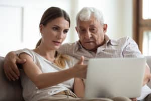 Senior father sits on couch with his daughter as they look at a laptop.