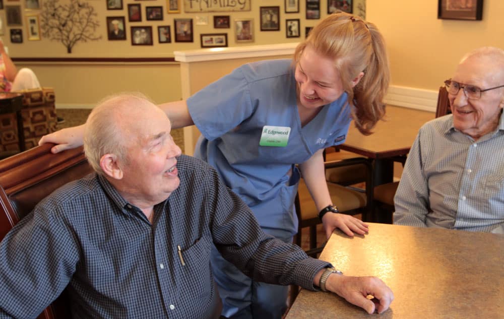 A young female nurse chats with two senior men in a dining room at Edgewood Healthcare