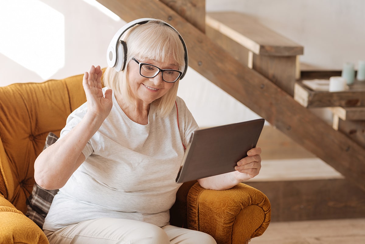 Female senior wearing white headphones waving at tablet screen