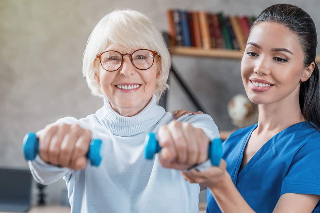 female senior lifting blue hand weights next to a female caregiver wearing blue scrub top