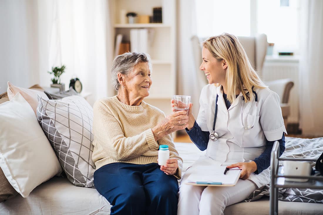 female senior wearing beige sweater, holding pills and receiving a glass of water from female health care professional holding clip board