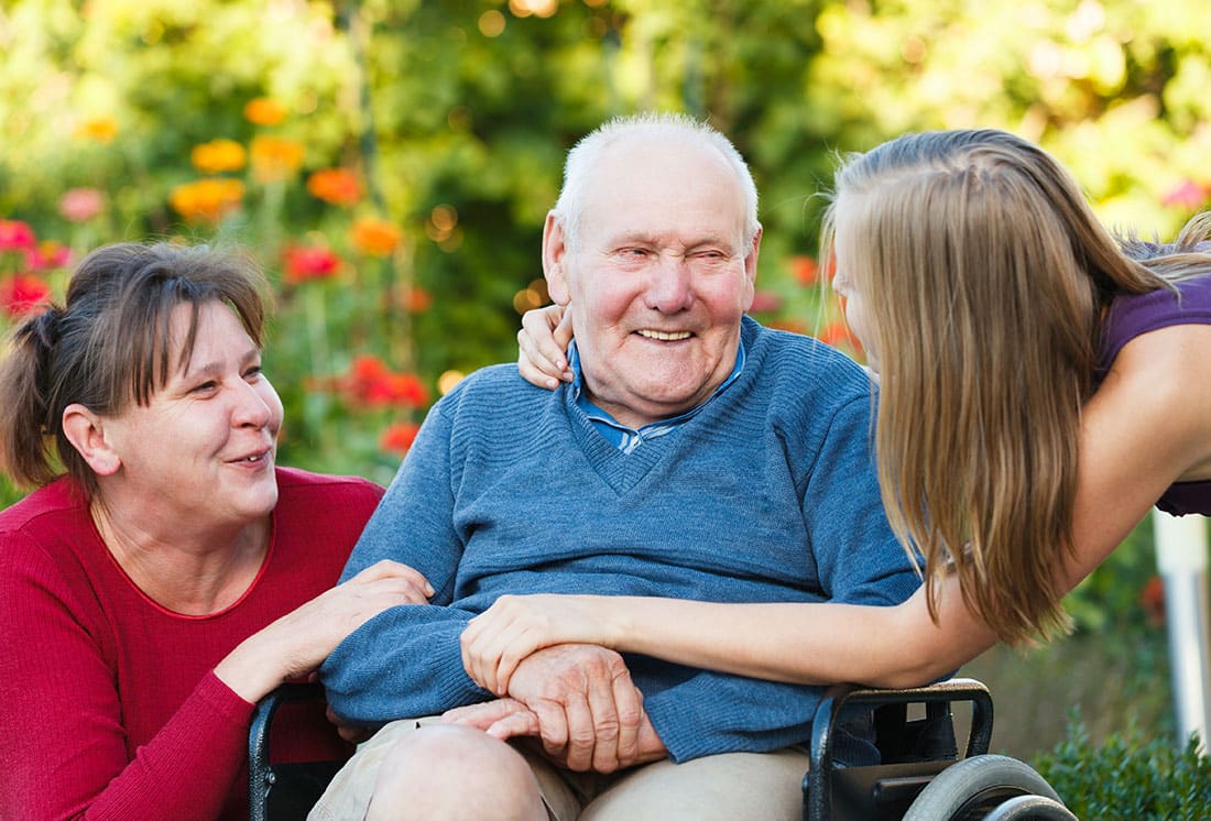 male senior wearing blue seated in wheelchair smiling with female loved one wearing red and female loved one reaching out to embrace