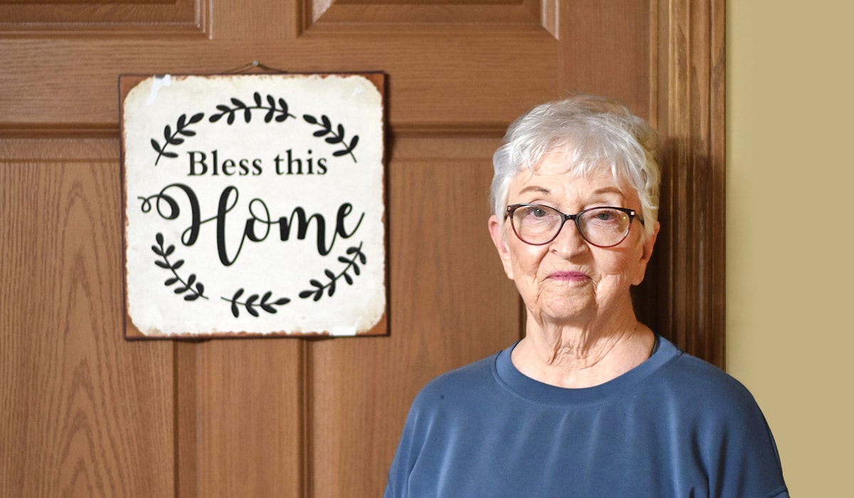 senior female wearing blue sweater next to a door sign that says “bless this home.”