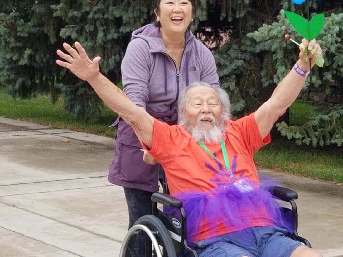 Man wearing an orange t-shirt with arms in the air being pushed in a wheelchair by female