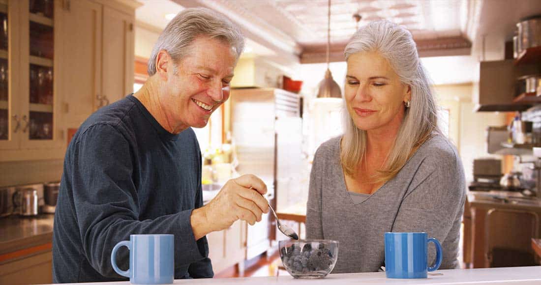 Senior male holding spoon near dish of blueberries seated next to senior female.
