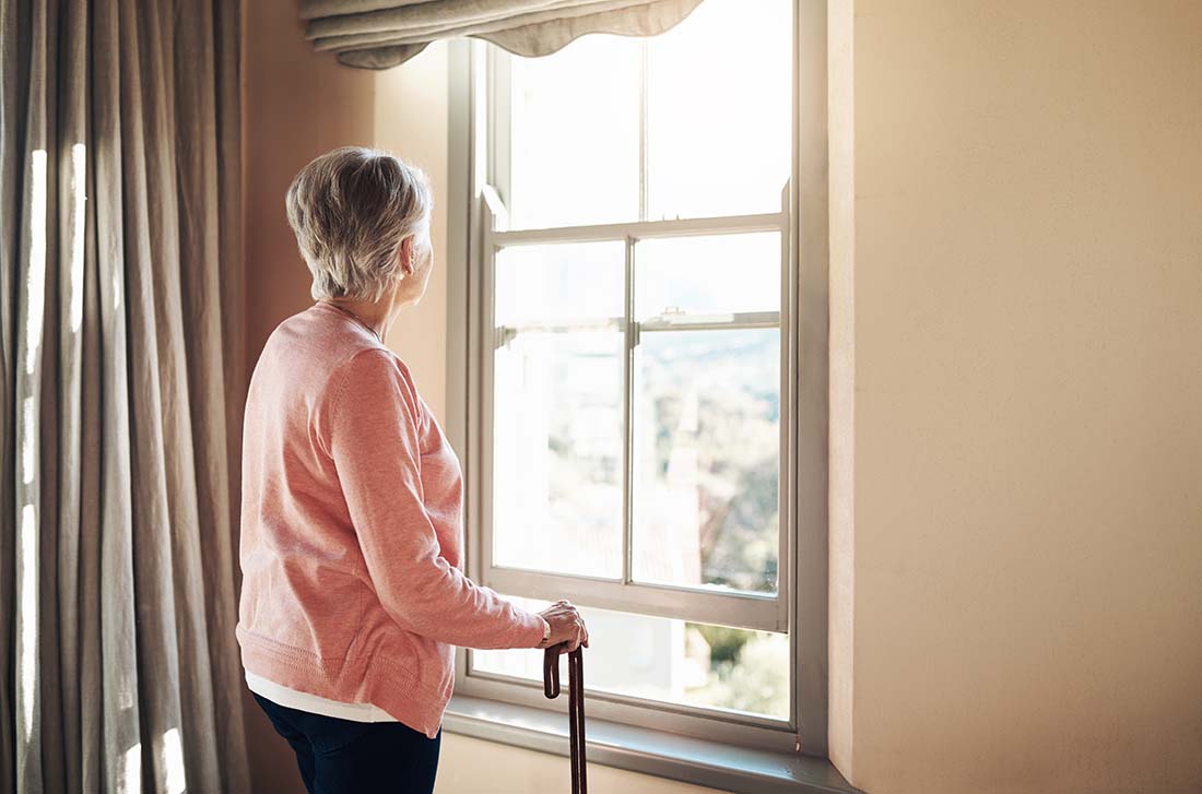 Senior female wearing pink holding a cane and looking out the window