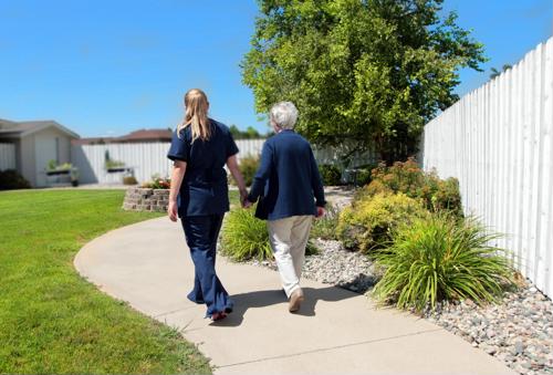 Female caregiver wearing blue scrubs holding hand of female senior walking outside