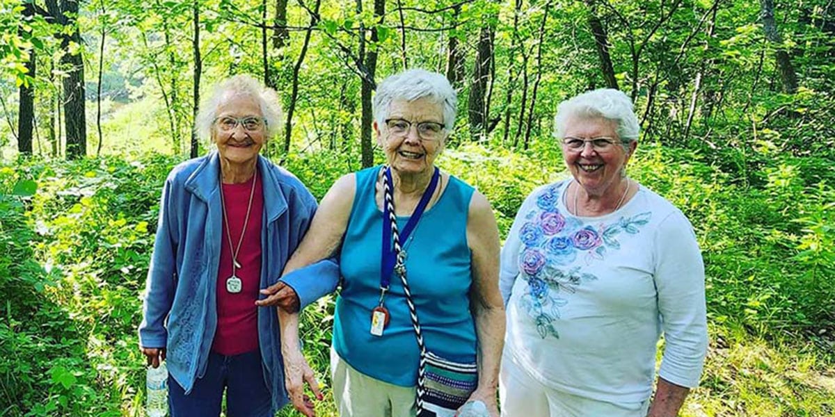 Three senior females standing outdoors in front of green bushes and trees.