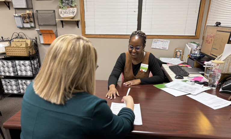 a woman sitting behind a desk showing another woman where to sign on papers