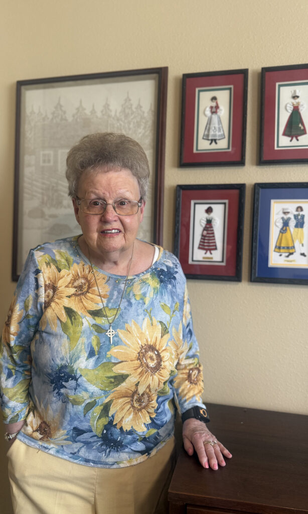 Older woman smiling while seated indoors with framed artwork displayed on the wall behind her.