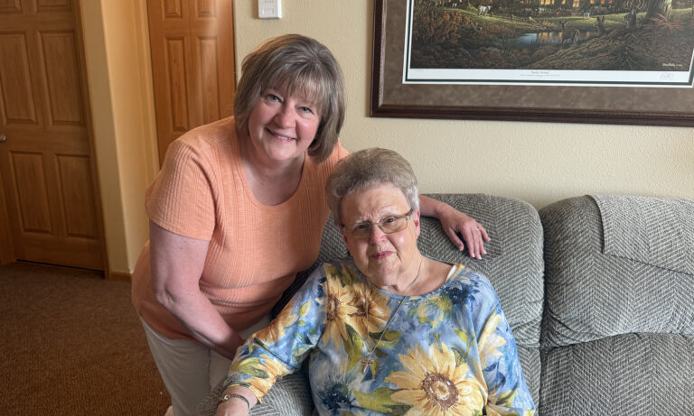 Older woman and her adult daughter smiling together on a couch in their home.