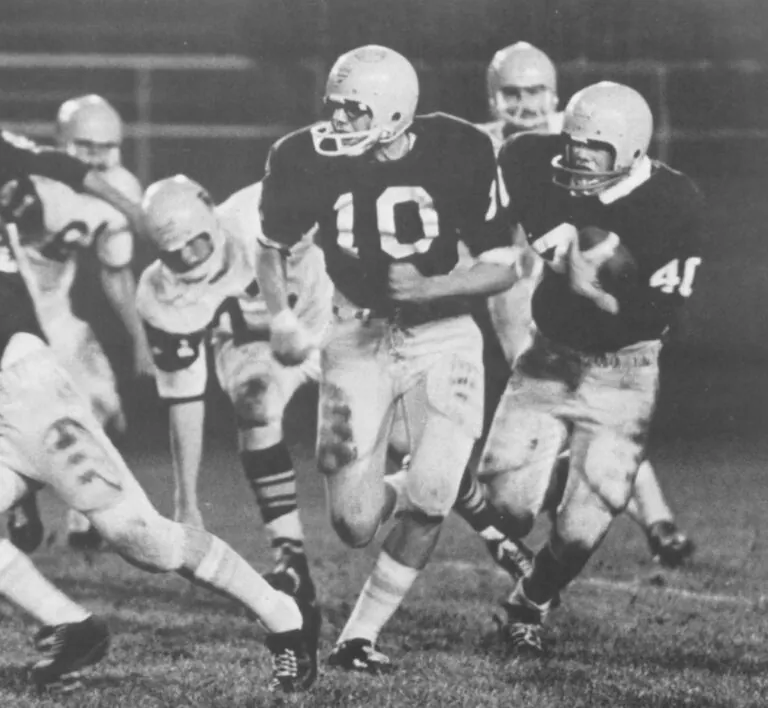 Black-and-white archival photo of a football game. A player wearing number 19 runs through a cluster of helmeted players as an opponent reaches toward him.