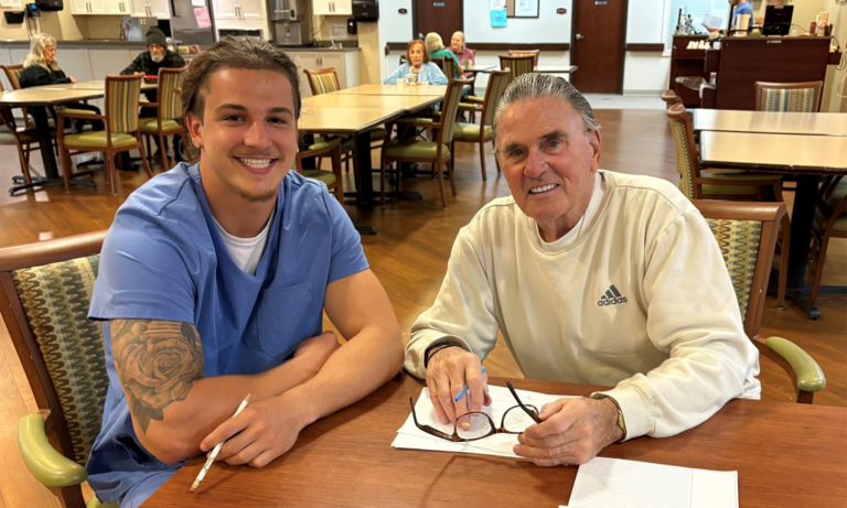 Joe Glenn, a memory care resident, smiles beside an Edgewood staff member in blue scrubs as they sit together over papers at a dining room table.