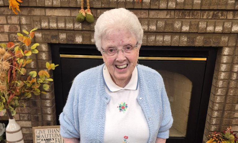An older woman with short white hair, glasses, and a light blue cardigan, standing in front of a brick fireplace decorated for fall.