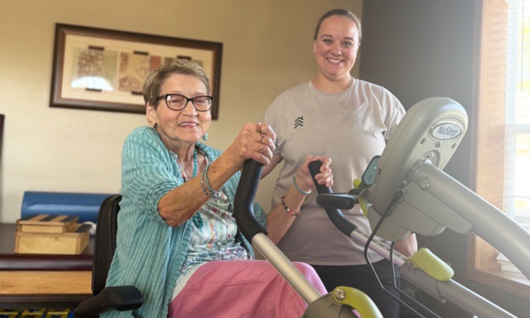 A senior woman holding the handlebars of a therapy machine while a female CaringEdge therapist stands closely behind her, assisting with the exercise.
